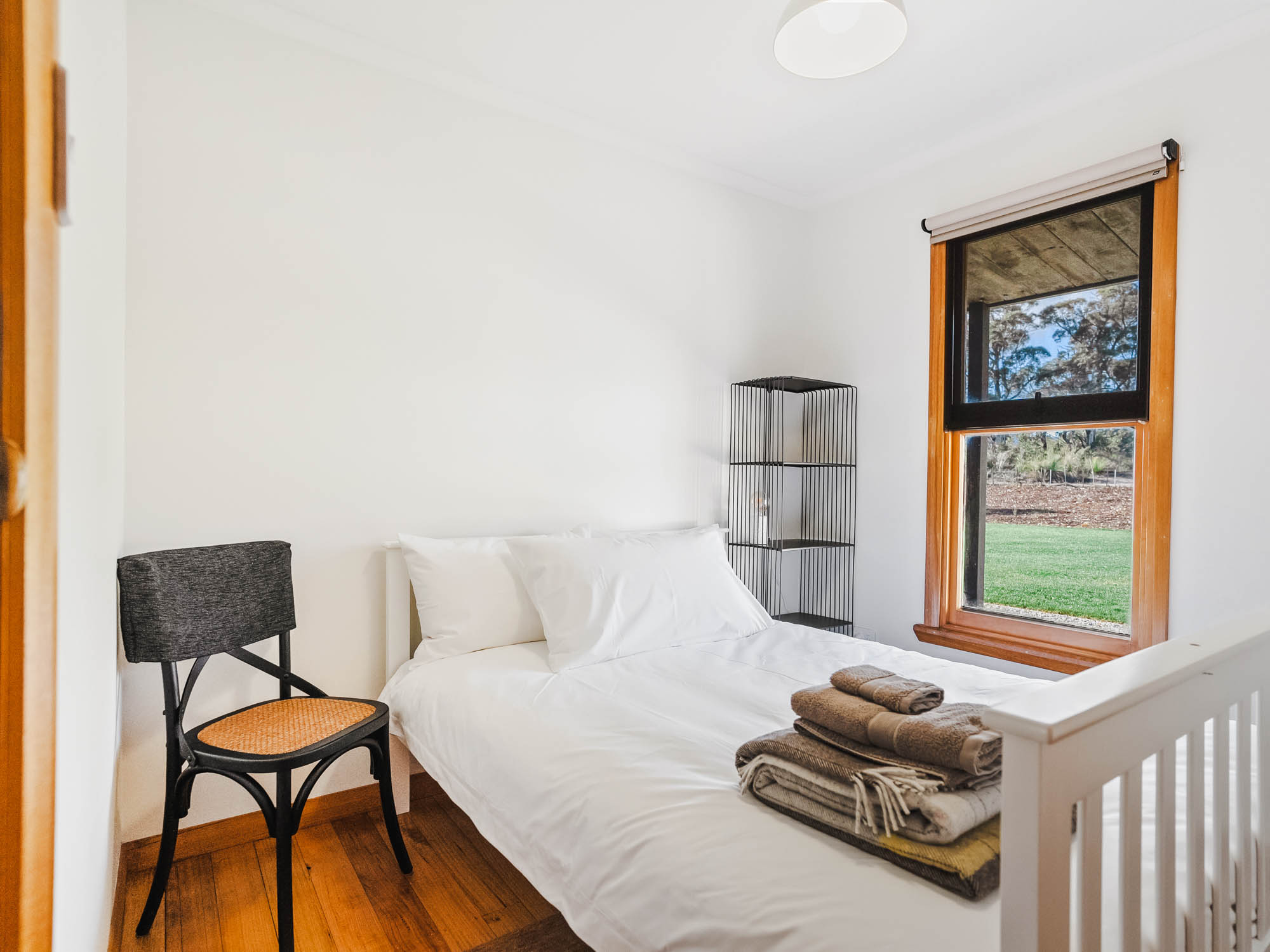 Minimalist bedroom with a white bed, neatly folded towels on top, a black chair, and a window showing green grass outside.