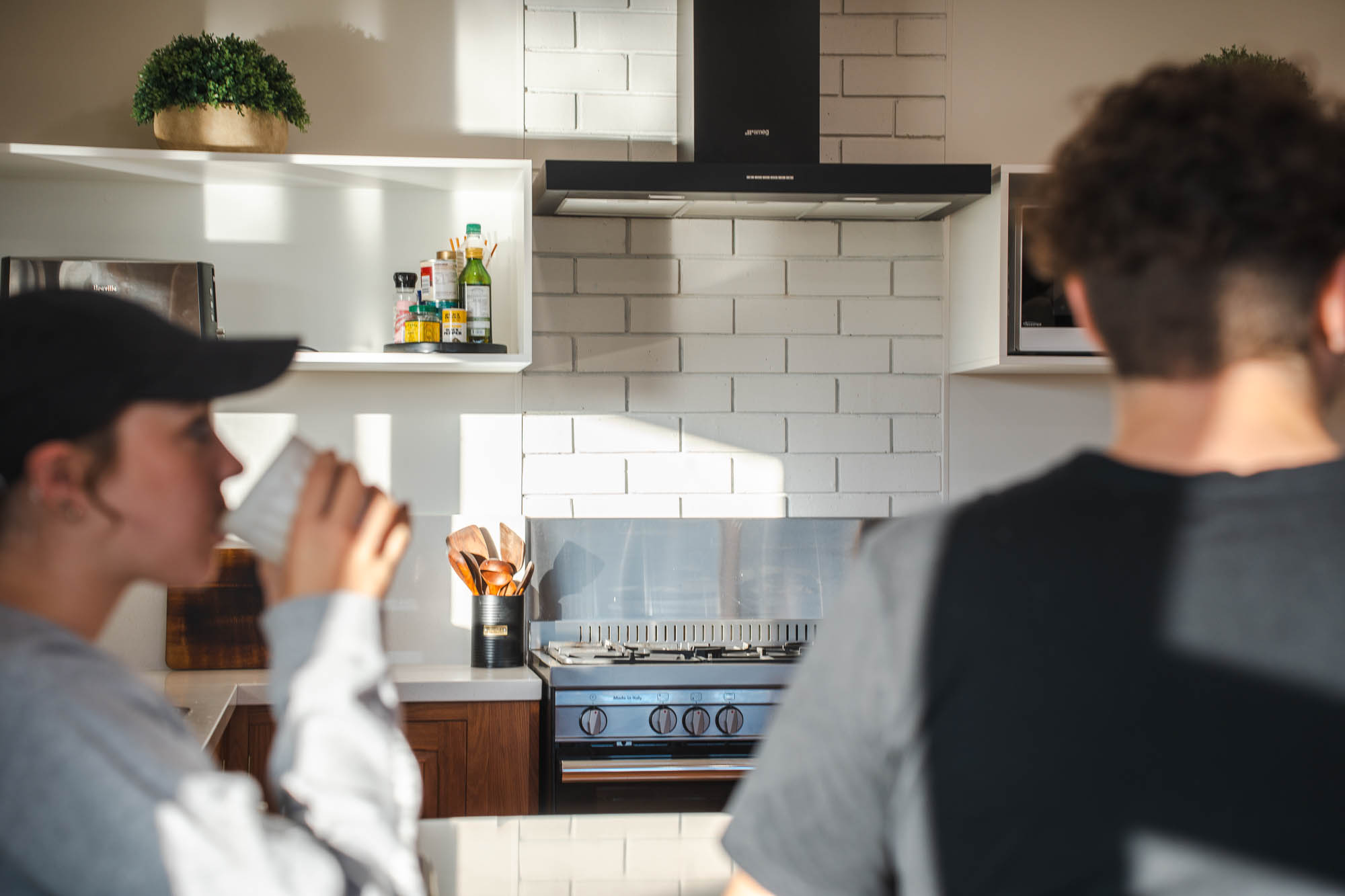 Two people in a kitchen, one drinking from a cup and the other facing away, with a stove, utensils, and kitchen shelves in the background.