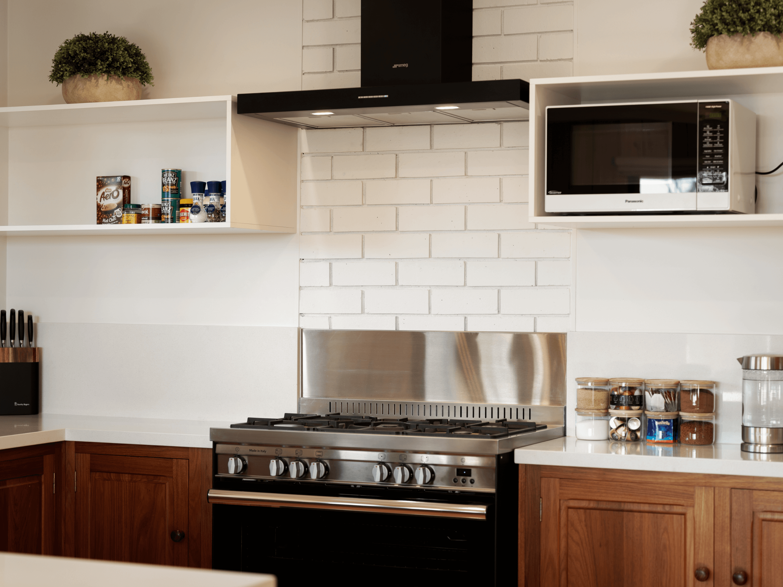 Modern kitchen with stainless steel gas stove, black range hood, microwave, wooden cabinets, spice jars, and plants on shelves.