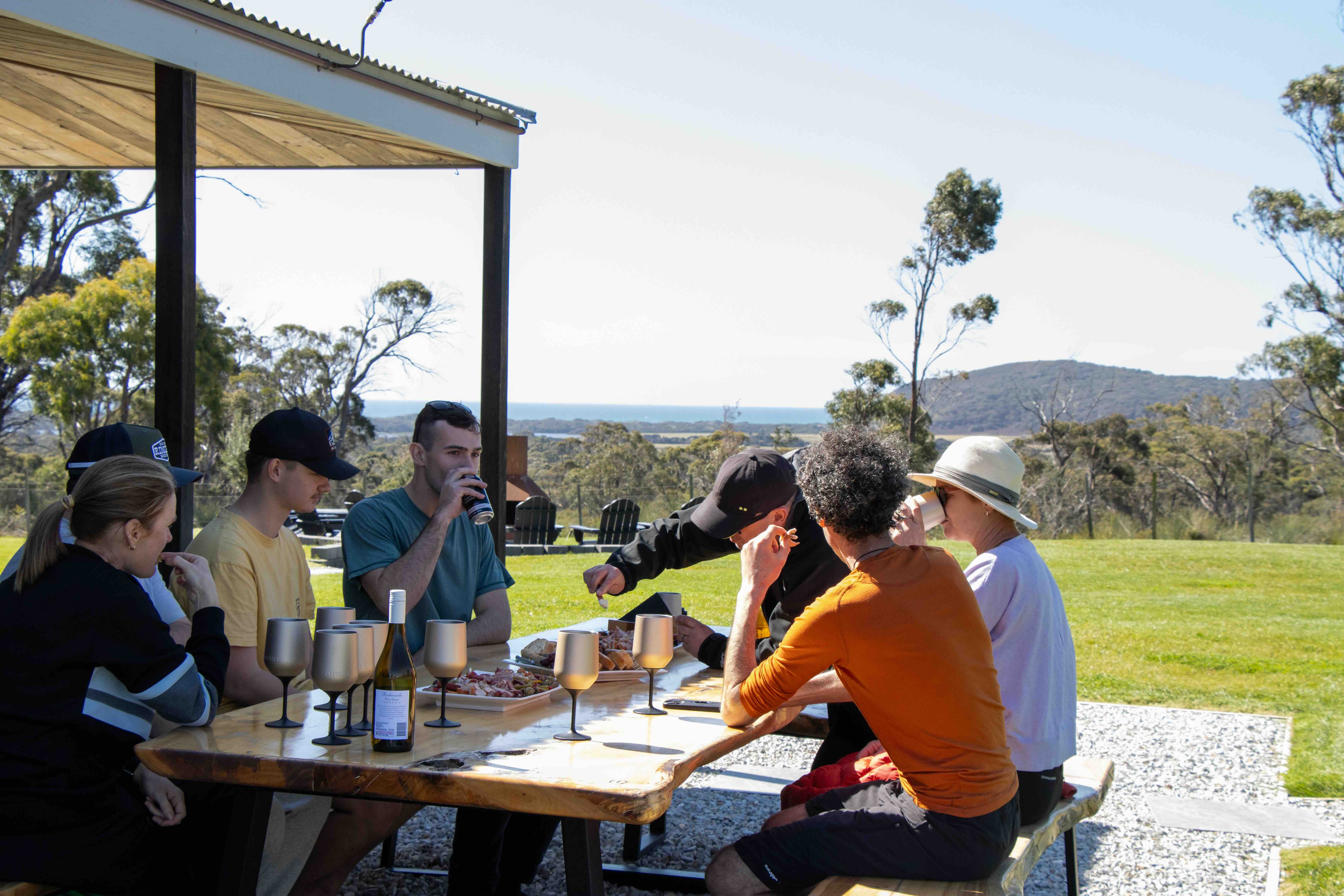 Group of six people sitting outdoors at a wooden picnic table with food and drinks, surrounded by green grass and trees under a clear sky.