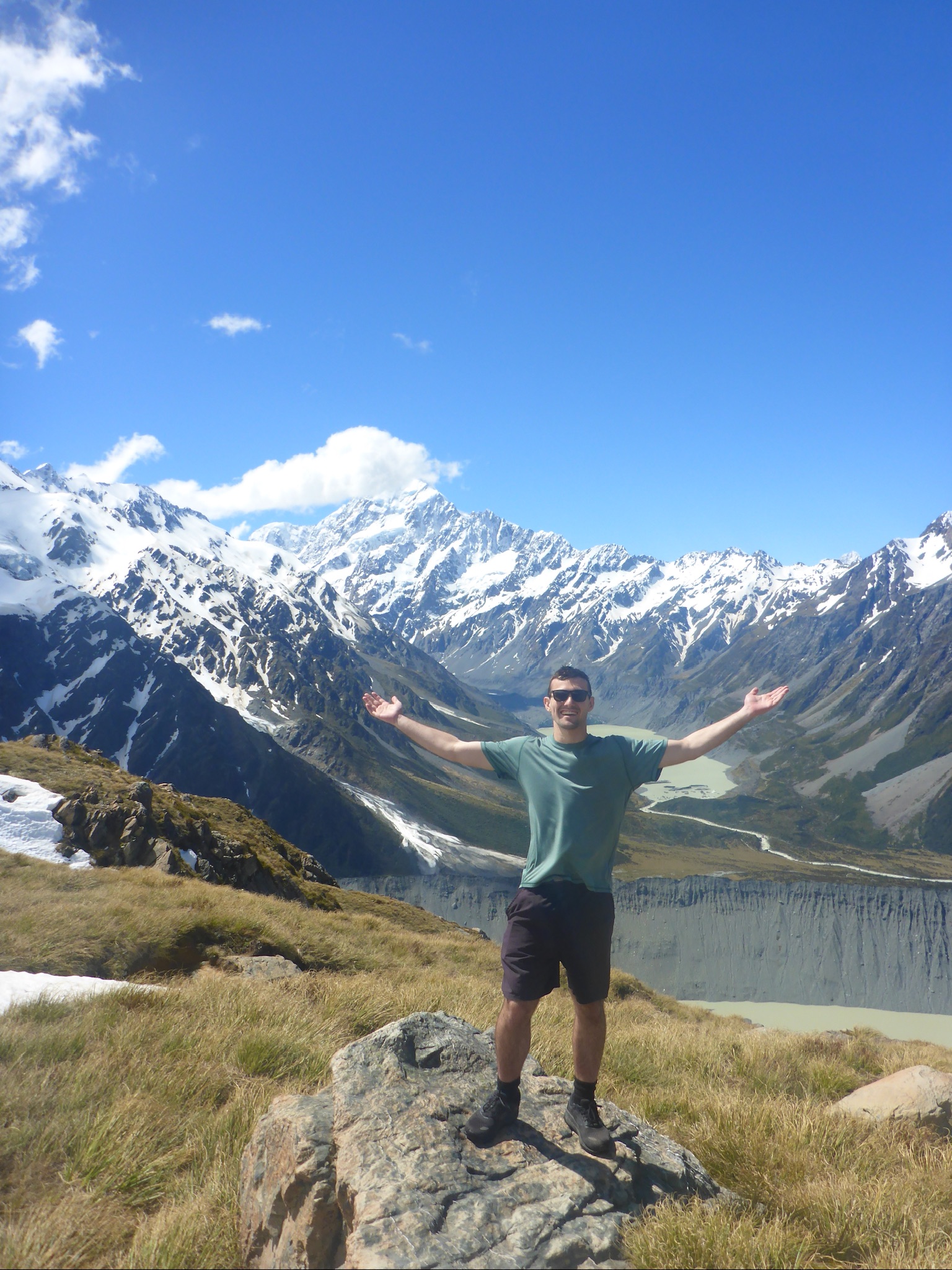 Man wearing sunglasses and green T-shirt standing on a rock with arms outstretched in a mountainous landscape with snow-capped peaks under a clear blue sky.