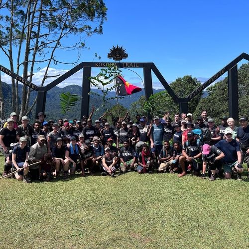 Large diverse group of hikers posing under a Kokoda Trail entrance arch with mountains in the background and a flag held up in the center.
