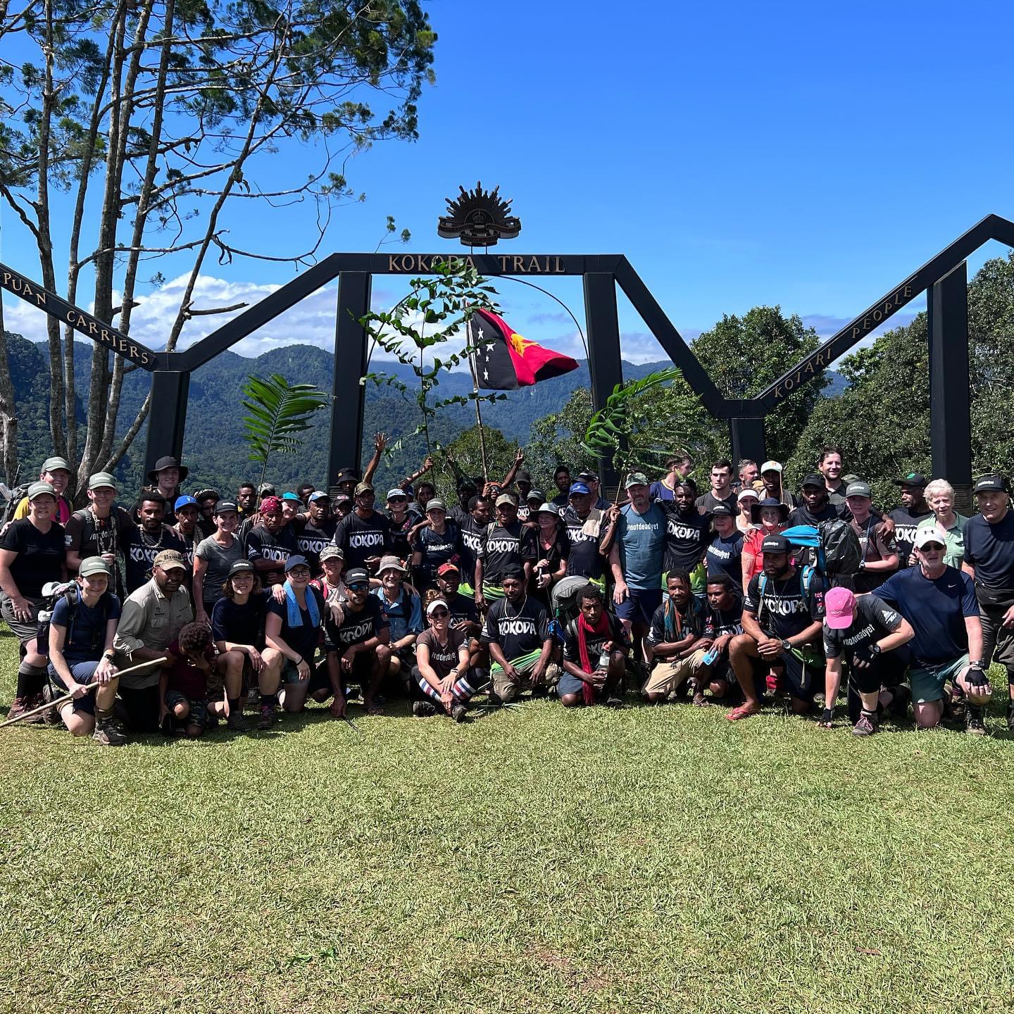 Large diverse group of hikers posing under a Kokoda Trail entrance arch with mountains in the background and a flag held up in the center.