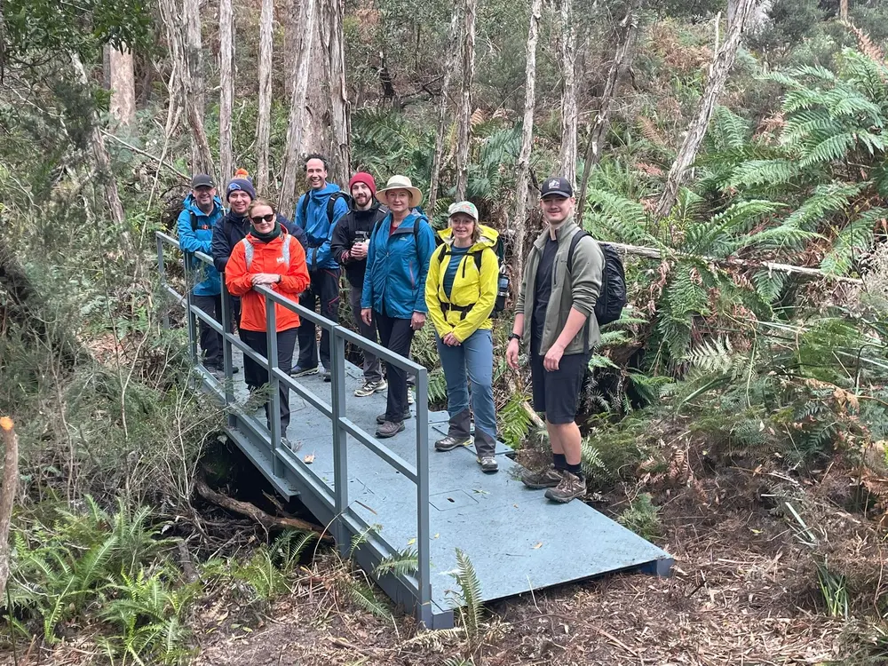 Group of seven hikers standing on a metal bridge in a forested area with tall trees and ferns.