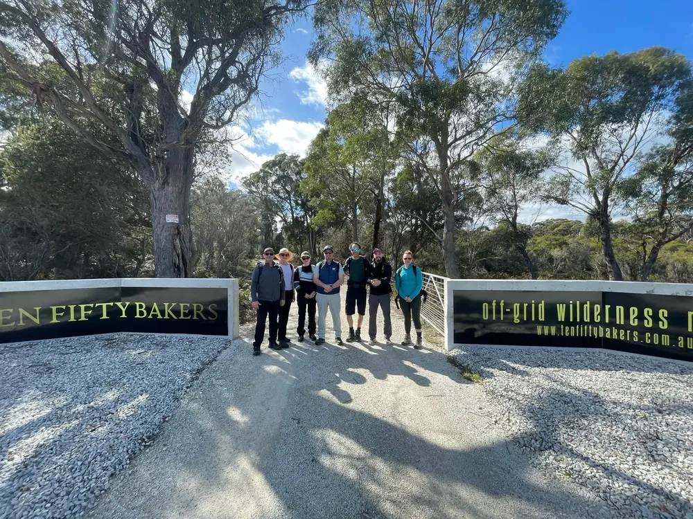 Group of six people standing on a gravel road between two signs reading 'Ten Fifty Bakers' and 'off-grid wilderness' at a forest entrance under a partly cloudy sky.