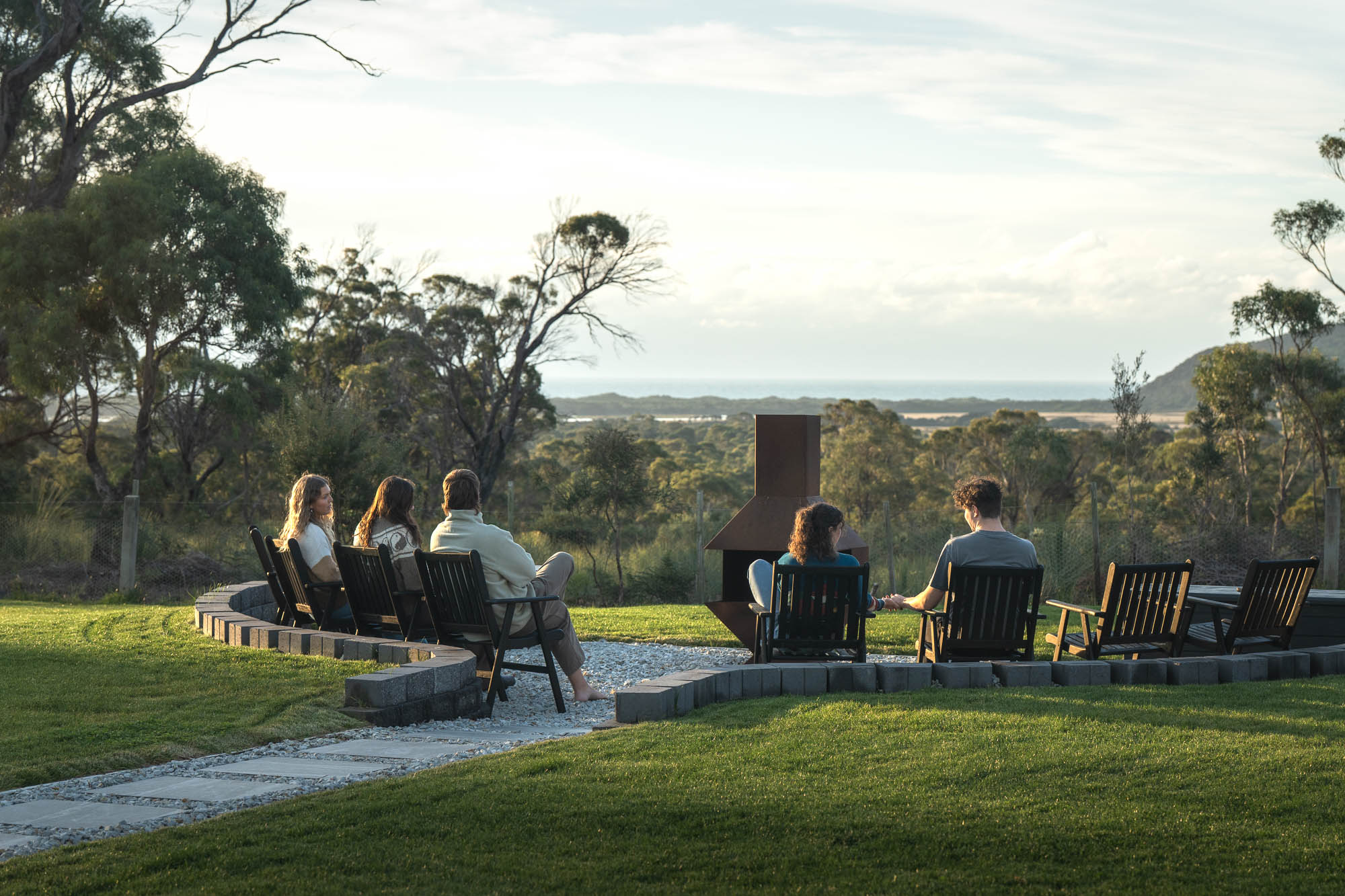Five people sitting on wooden chairs arranged in a semicircle around an outdoor fireplace in a grassy area with trees and hills in the background.