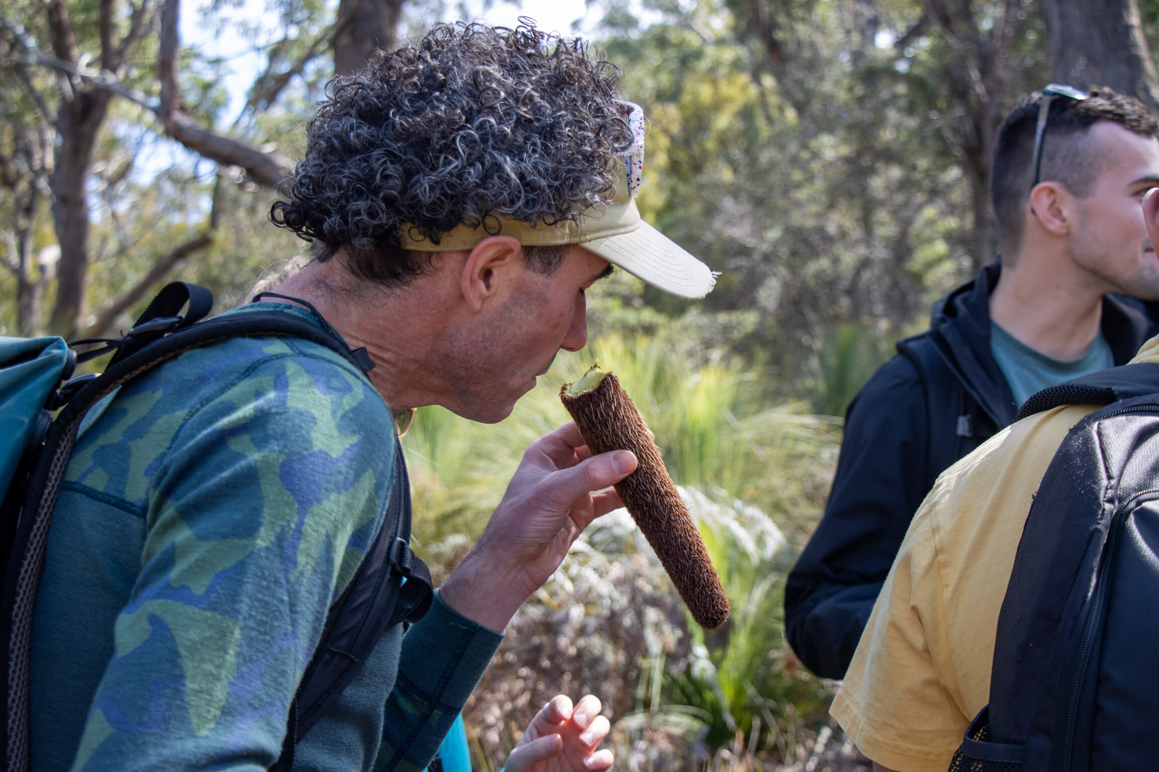 Man with curly hair and a beige cap eating a brown cylindrical plant pod outdoors.