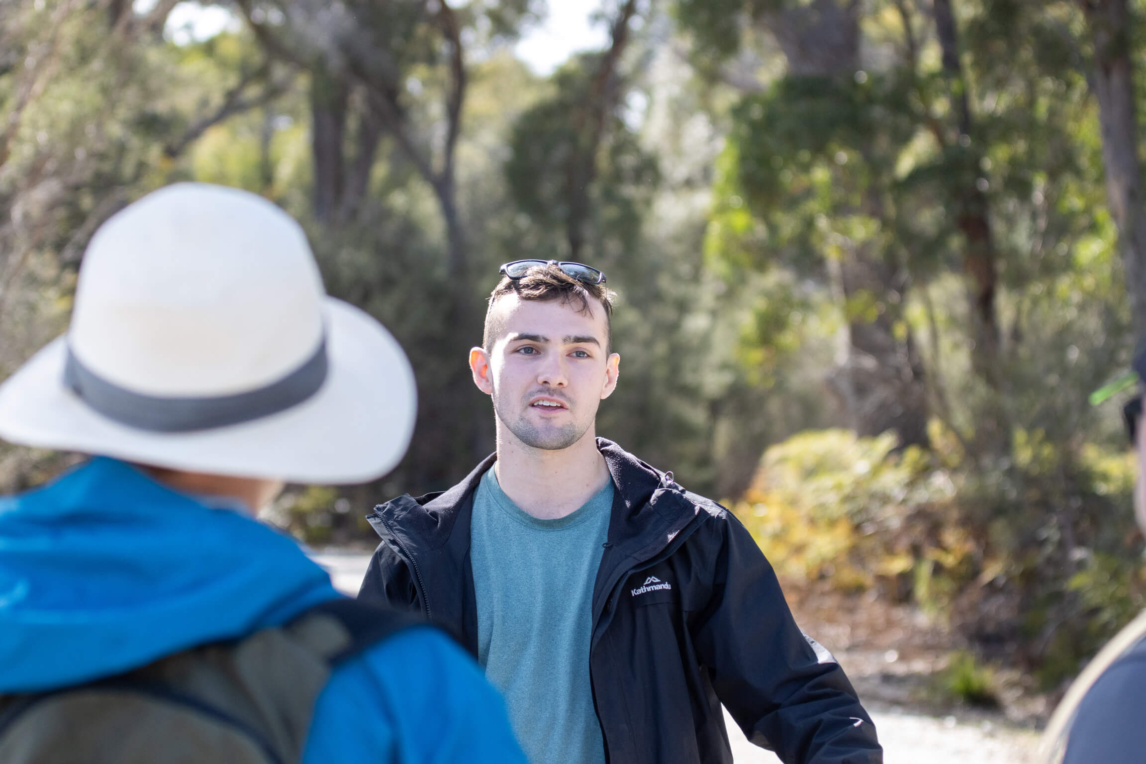 A group of eight people standing and listening to a guide speaking on a forest trail surrounded by tall trees and dry vegetation.