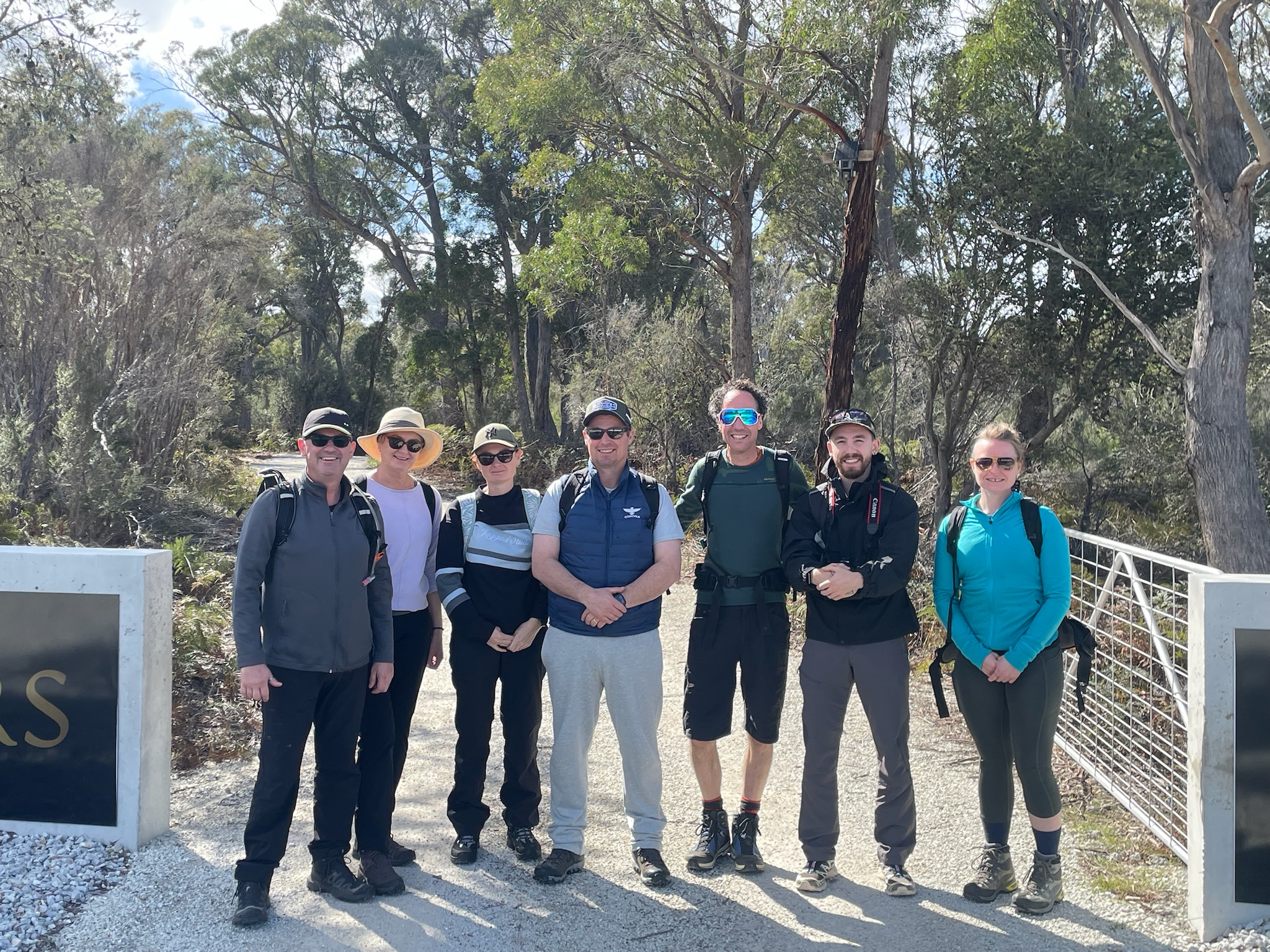 Eight hikers standing on a gravel path flanked by trees, dressed in outdoor clothing and carrying backpacks.