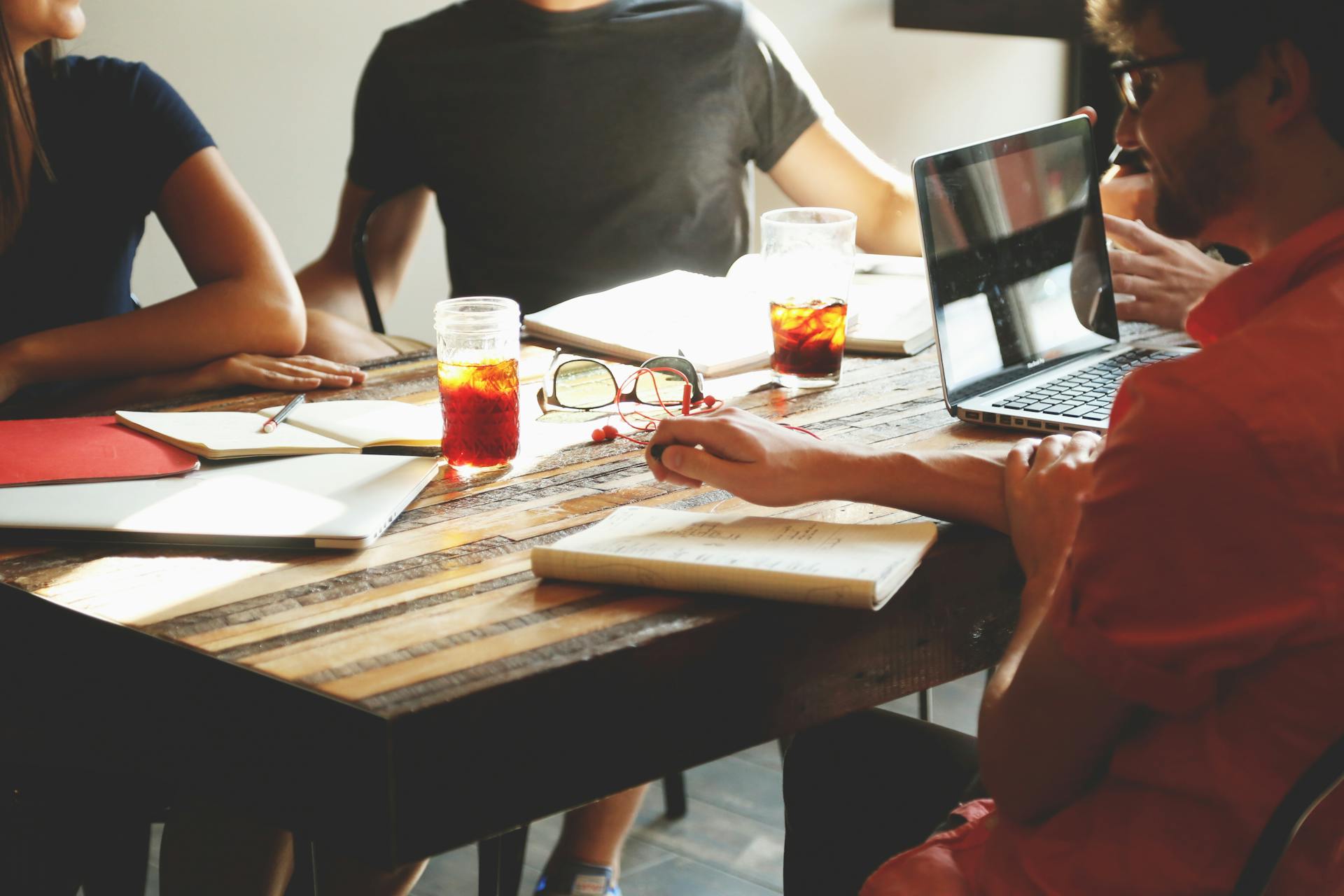 People sitting around a wooden table with notebooks, glasses of iced tea, a laptop, and reading glasses.