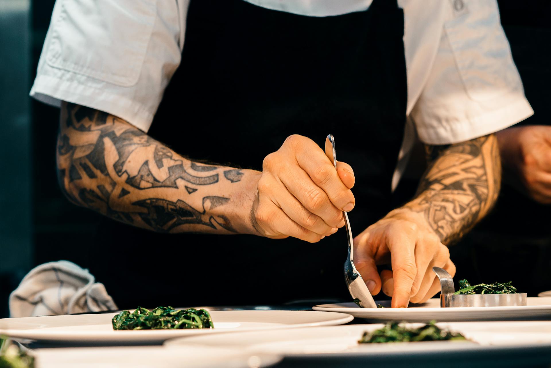 Chef with tattooed arms plating a dish of cooked greens on white plates.