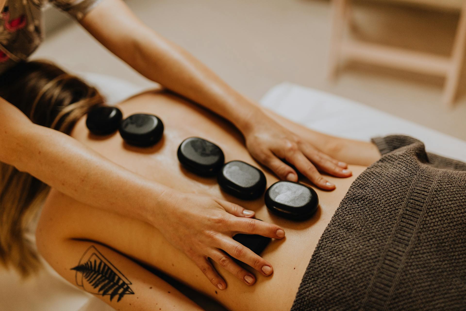 Person receiving hot stone massage with five black stones placed on their back and hands of therapist applying pressure.