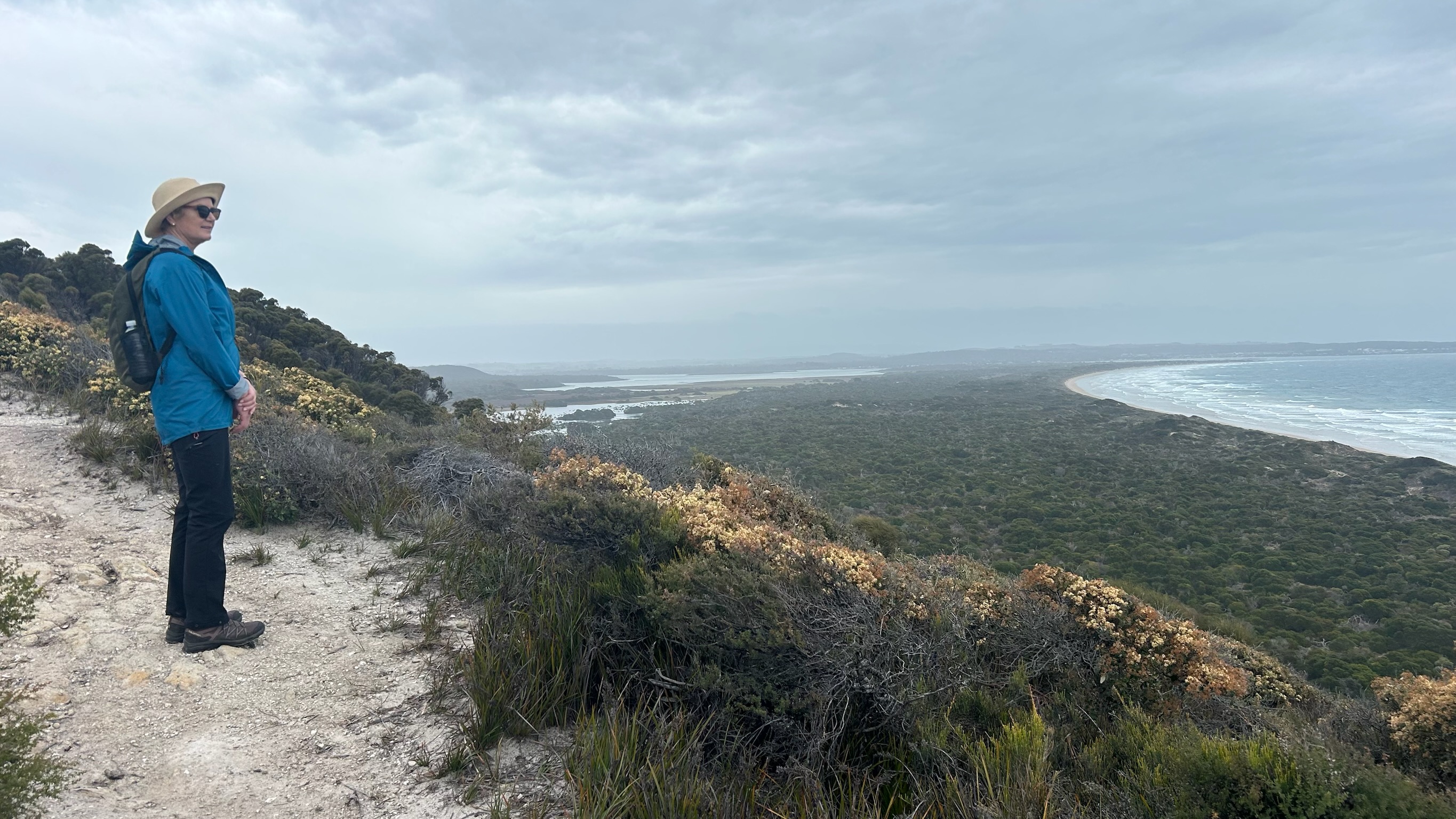 Person wearing a blue jacket and hat standing on a sandy trail overlooking a coastal landscape with dense greenery and a curved beach under a cloudy sky.